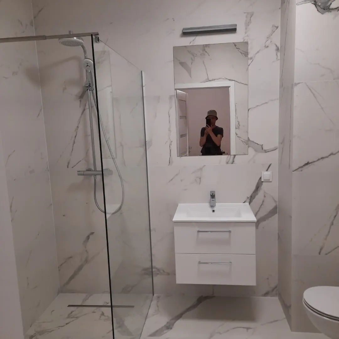 Nowoczesna łazienka z kabiną prysznicową typu walk-in, białą szafką z umywalką i lustrem, wykończona płytkami imitującymi marmur, z widocznym odbiciem osoby robiącej zdjęcie w lustrze.