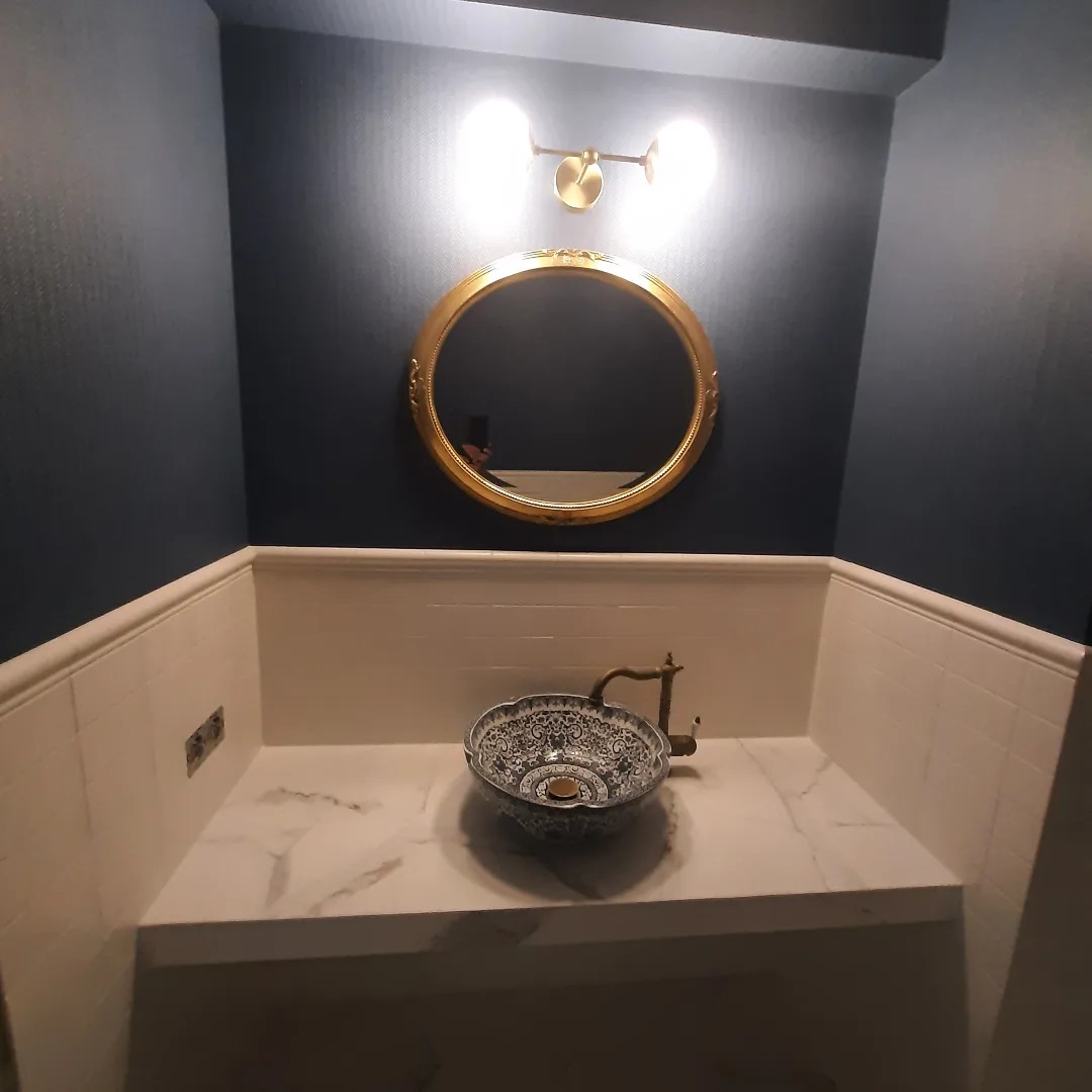 Dekoracyjna umywalka nablatowa w kolorze niebiesko-białym, z mosiężną armaturą, osadzona na blacie z białego marmuru w łazience z granatowymi ścianami i okrągłym lustrem w złotej ramie.