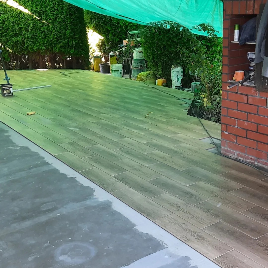 Układanie podłogi z płytek imitujących drewno na zewnątrz w Warszawie, widoczne narzędzia glazurnicze i zaplecze z zielenią.