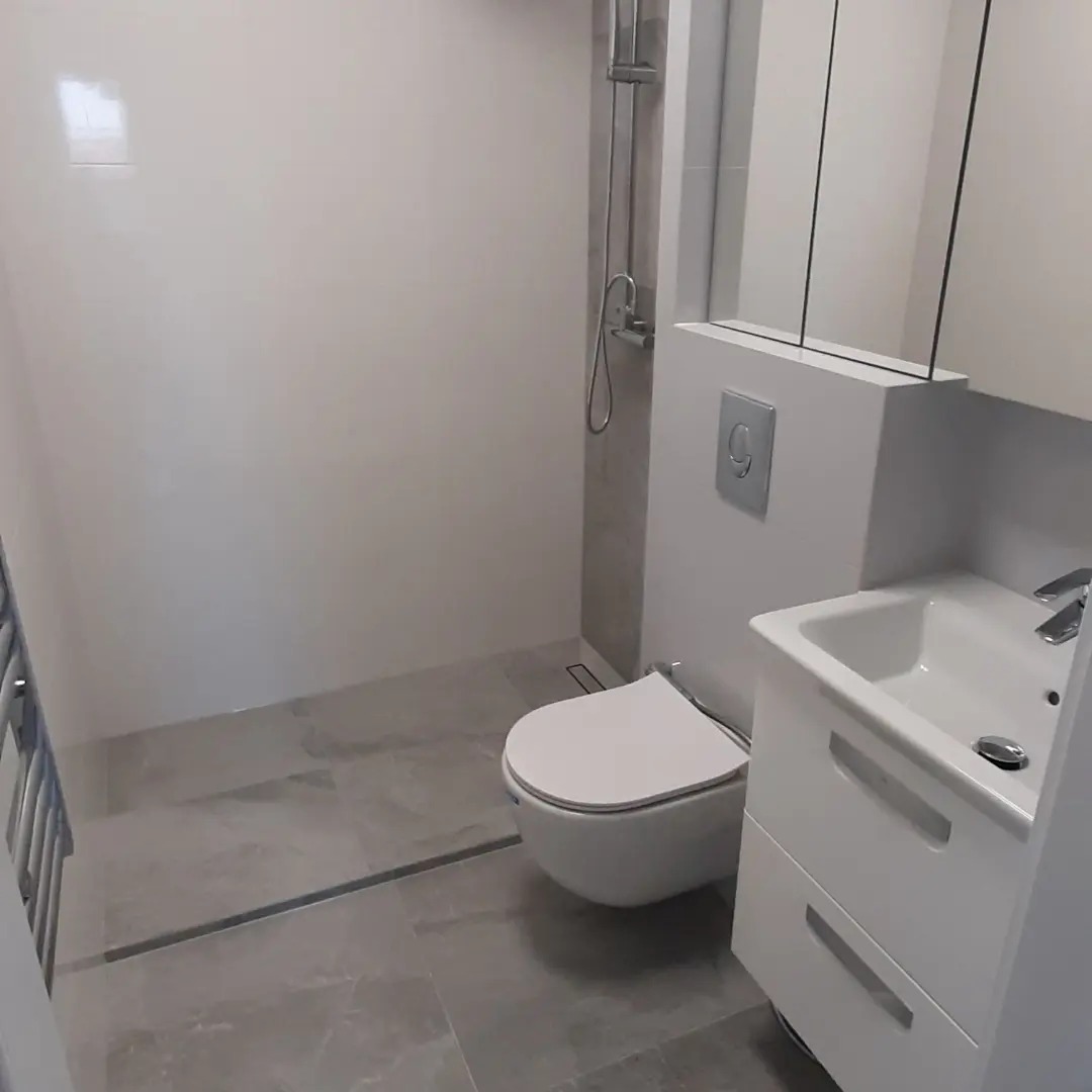 Nowoczesna łazienka w szarościach z podwieszaną toaletą, kabiną prysznicową typu walk-in i białą szafką umywalkową z dwiema szufladami.