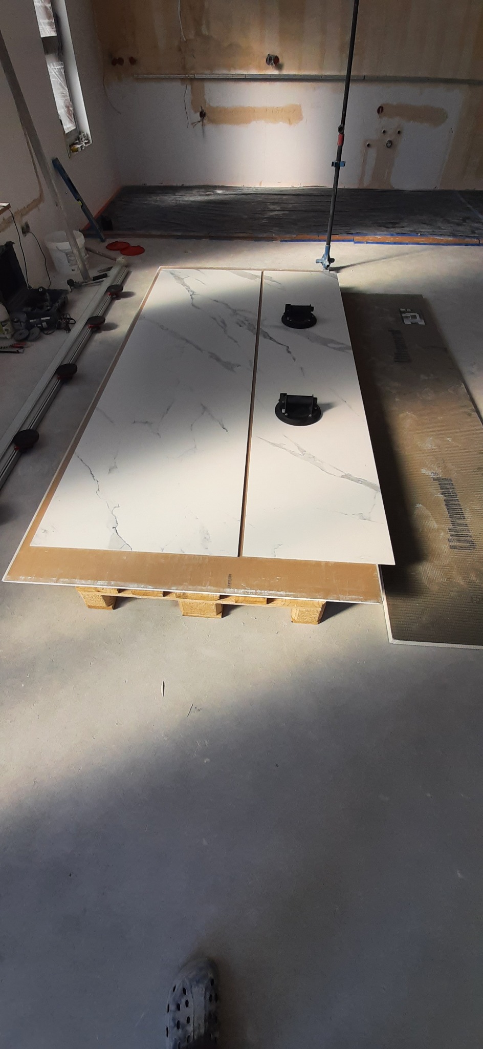 Dwie płyty wielkoformatowe z wzorem marmuru, podparte na palecie, z przyssawkami do przenoszenia; w tle przygotowane podłoże pod posadzkę w trakcie remontu.