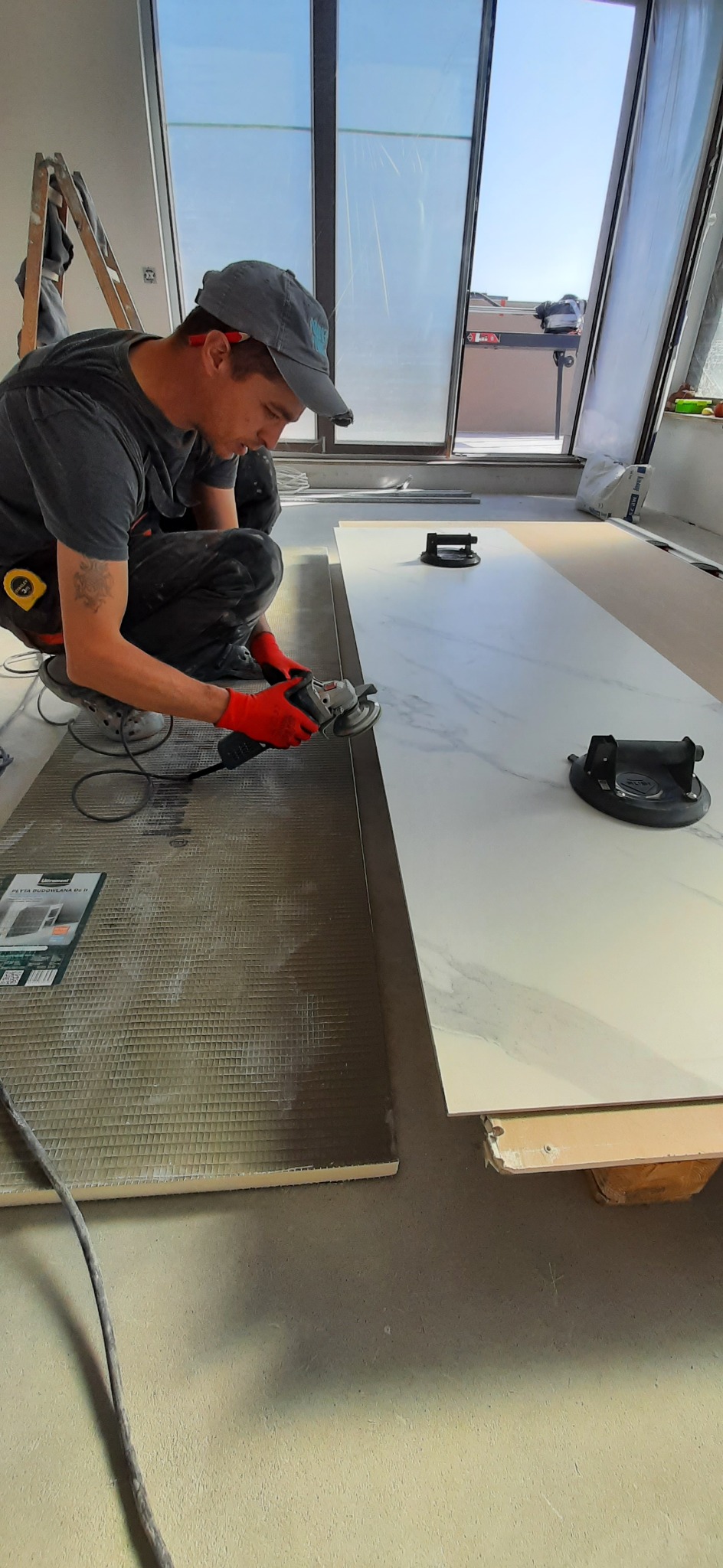 Pracownik w czerwonych rękawicach szlifuje krawędź płytki gresowej o wzorze marmuru szlifierką kątową, obok leży płyta budowlana do montażu pod płytki, widoczne przyssawki do przenoszenia płytek.