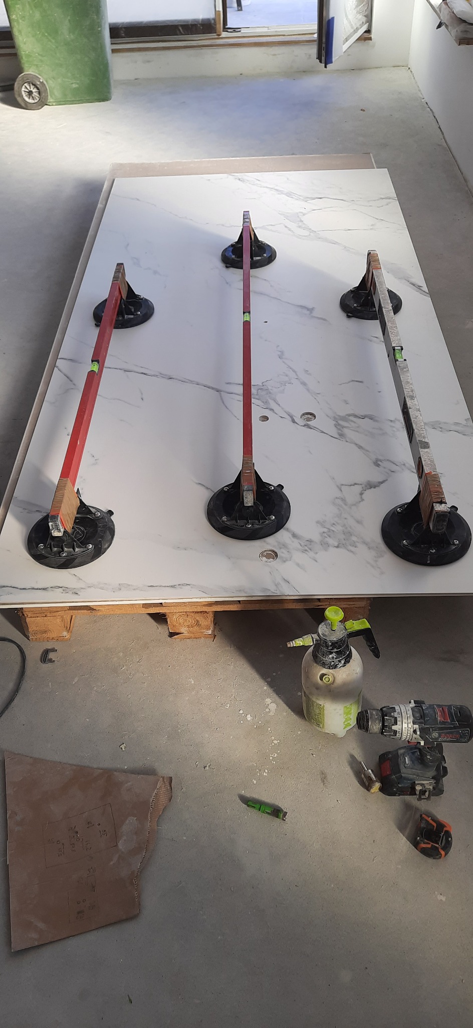 Biała płyta z marmurkowym wzorem, podtrzymywana przyssawkami z poziomicami, leżąca na palecie, obok narzędzia: wiertarka, spryskiwacz, nóż i laserowy dalmierz.