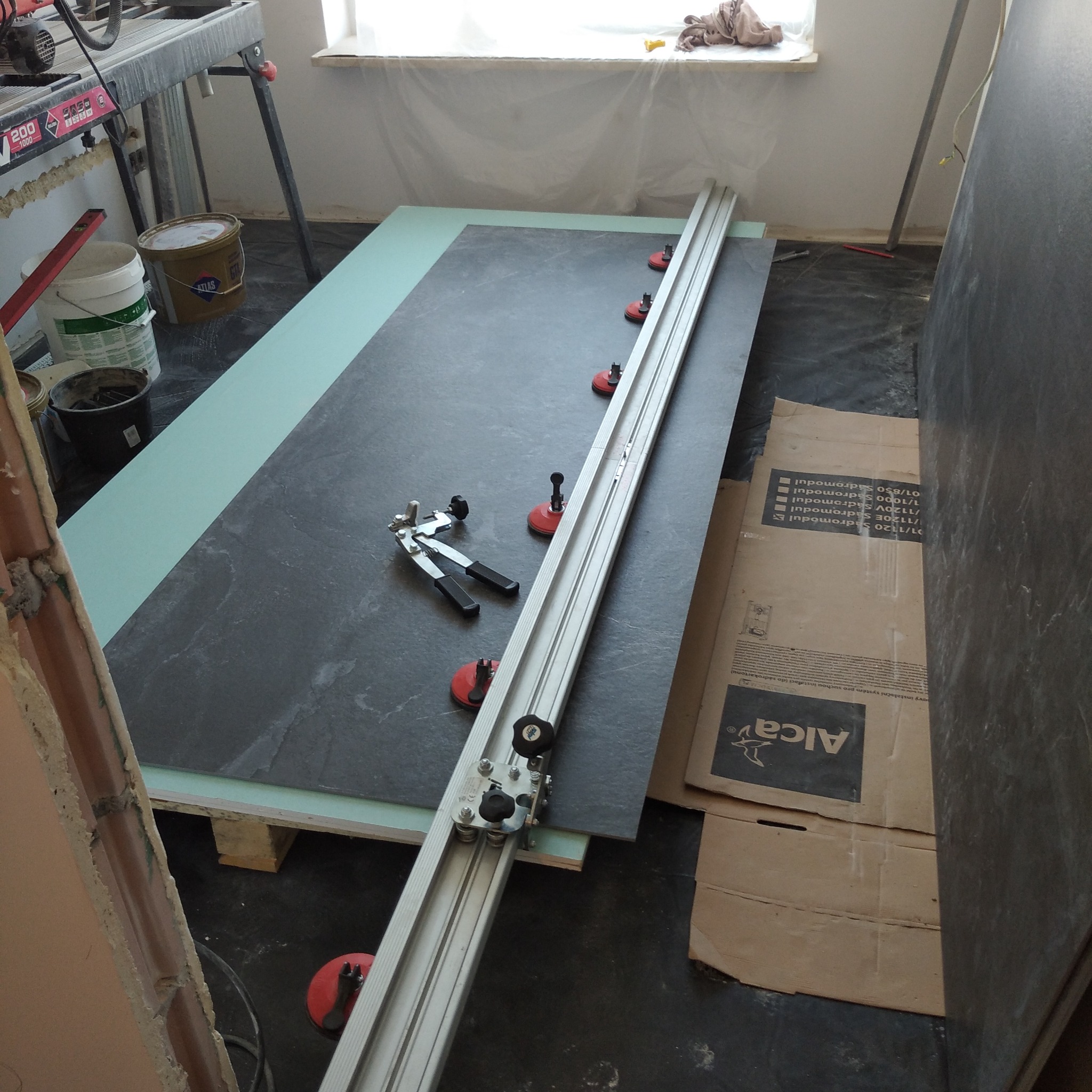 Przygotowanie do cięcia dużej, ciemnoszarej płytki ceramicznej za pomocą specjalistycznej przecinarki z przyssawkami, widoczne narzędzia i fragment ściany w trakcie prac.