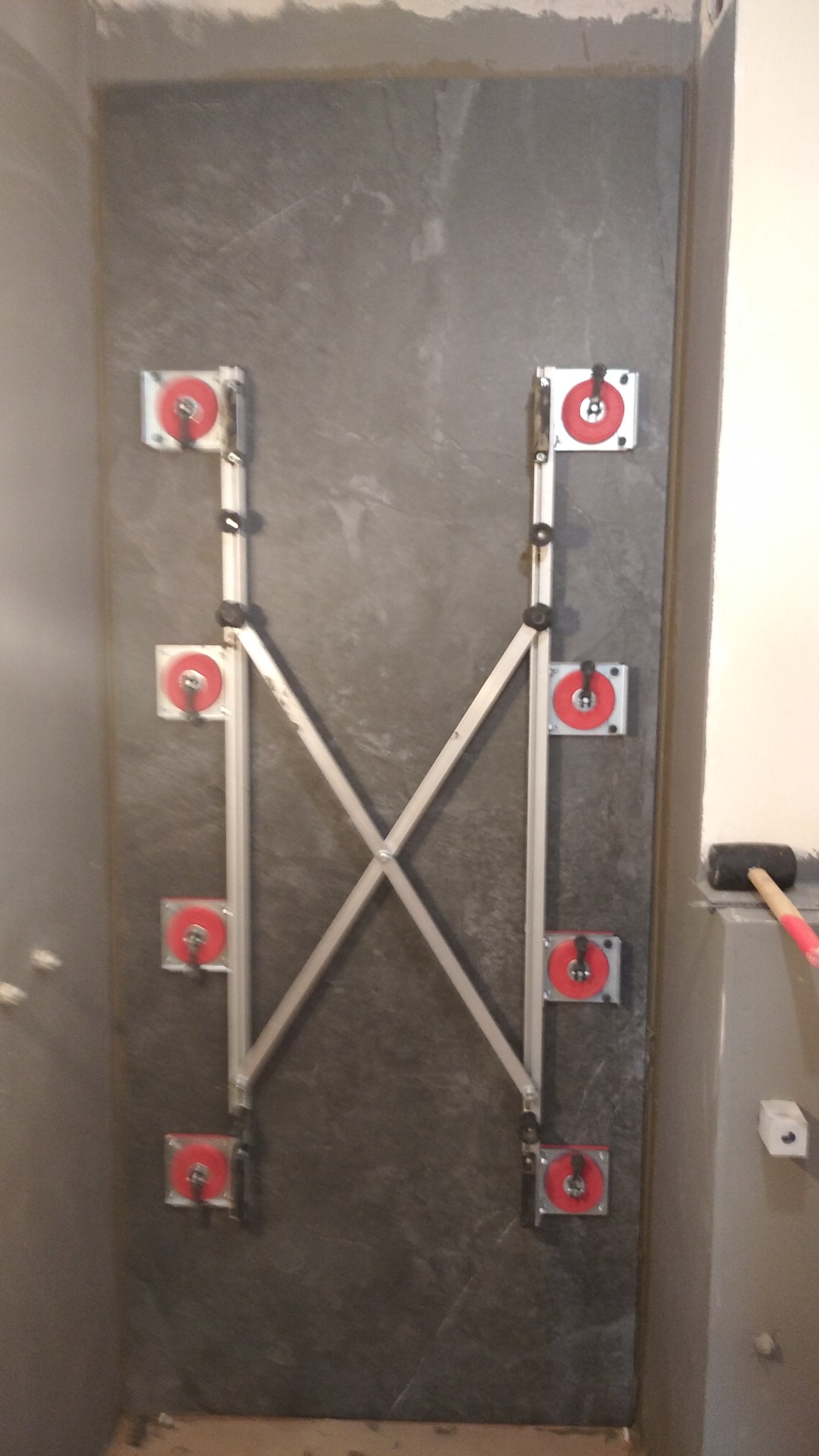 Szara płytka ścienna mocowana za pomocą aluminiowego systemu przyssawkowego z czerwonymi tarczami, młotek gumowy w tle.