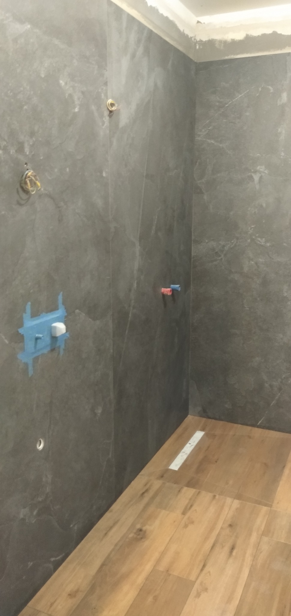 Szara, marmurkowa glazura ścienna w trakcie montażu w łazience, widoczne przygotowane instalacje wodne i elektryczne, oraz fragment podłogi wyłożonej panelami imitującymi drewno.