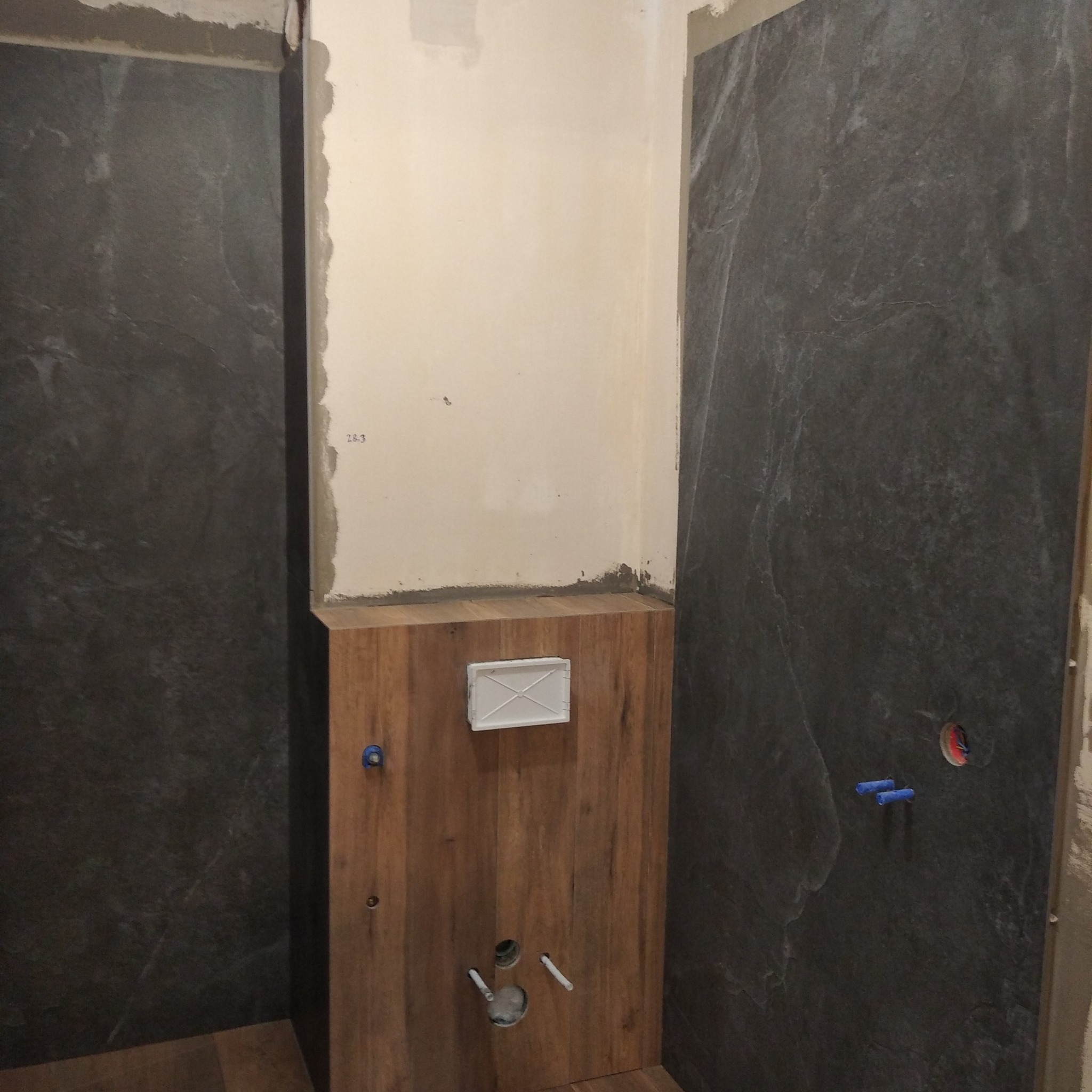 Łazienka w trakcie prac wykończeniowych: położone płytki imitujące kamień na ścianach, drewnopodobna zabudowa stelaża WC z zamontowanym przyciskiem, widoczne instalacje wodne i kanalizacyjne.