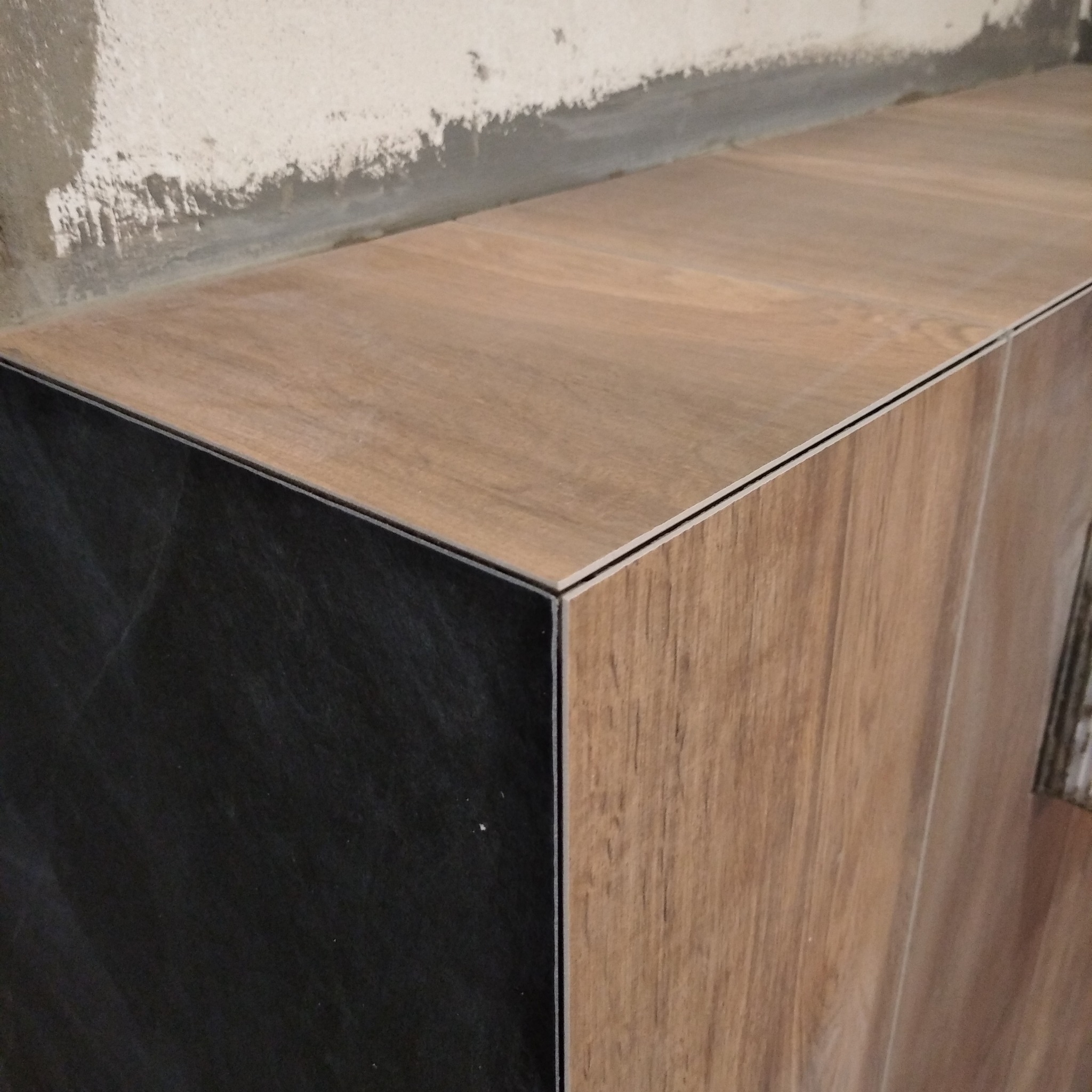 Wykończona obudowa z płytek imitujących drewno i czarny kamień, z widocznymi fugami i surową ścianą w tle.