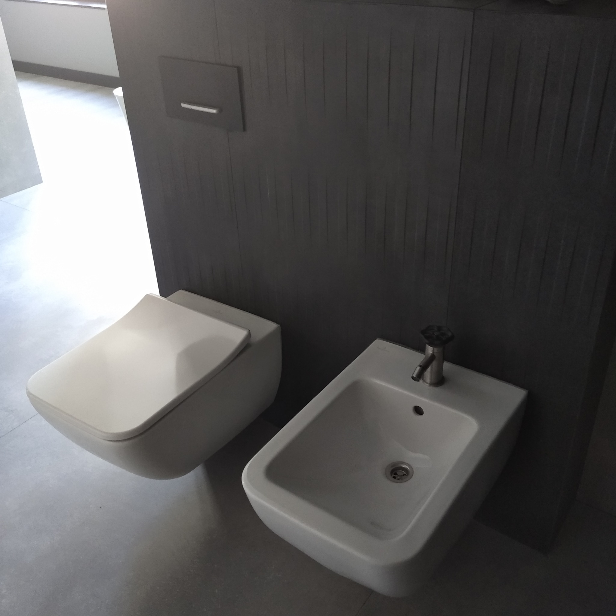 Nowoczesna łazienka z podwieszaną toaletą i bidetem, szara ściana z pionowymi panelami, minimalistyczny design.