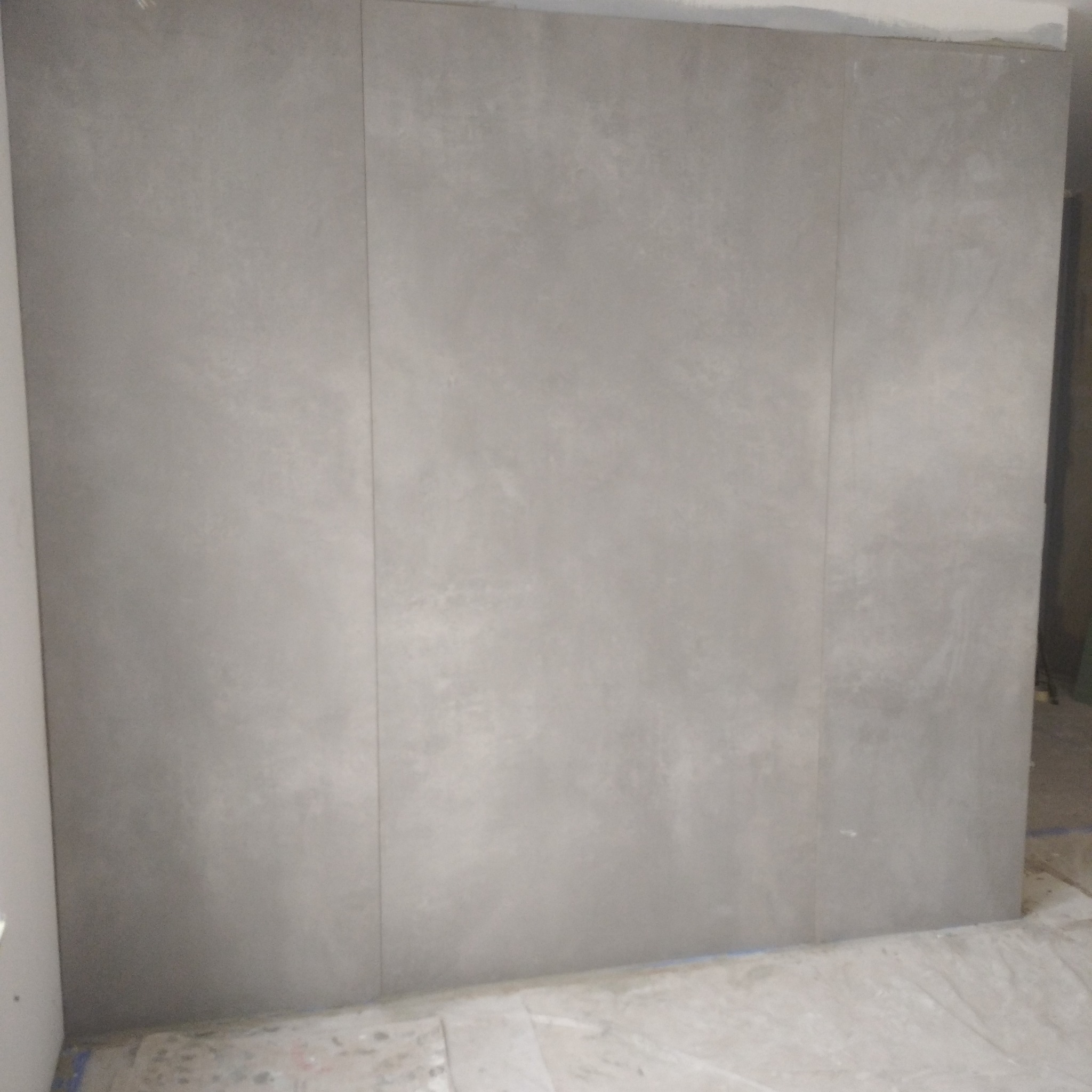 Ściana wyłożona płytami z szarego betonu architektonicznego, widoczne fugi między płytami, podłoga zabezpieczona folią malarską.