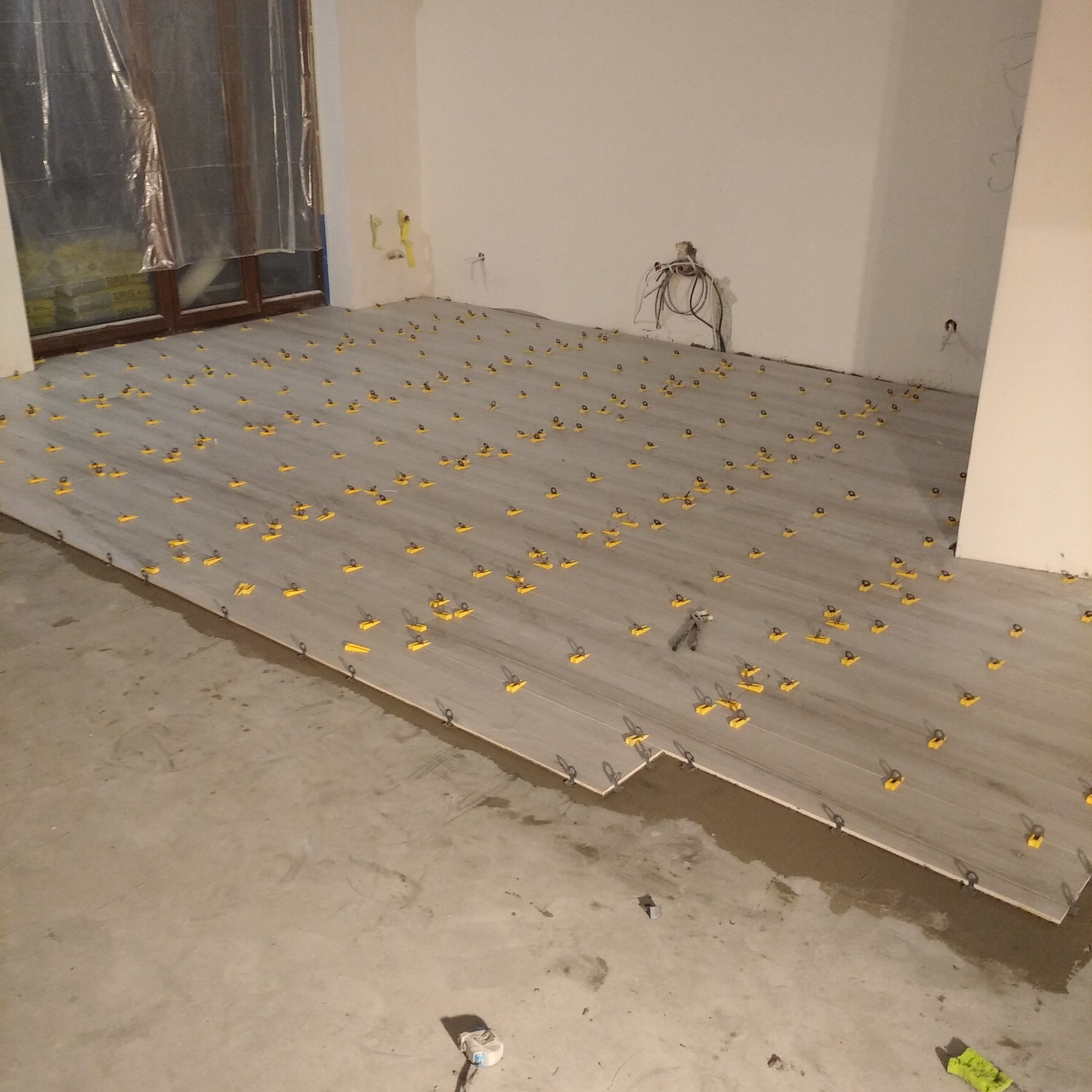 Układanie szarych płytek podłogowych z użyciem żółtych klipsów poziomujących, widoczne narzędzia i resztki zaprawy.