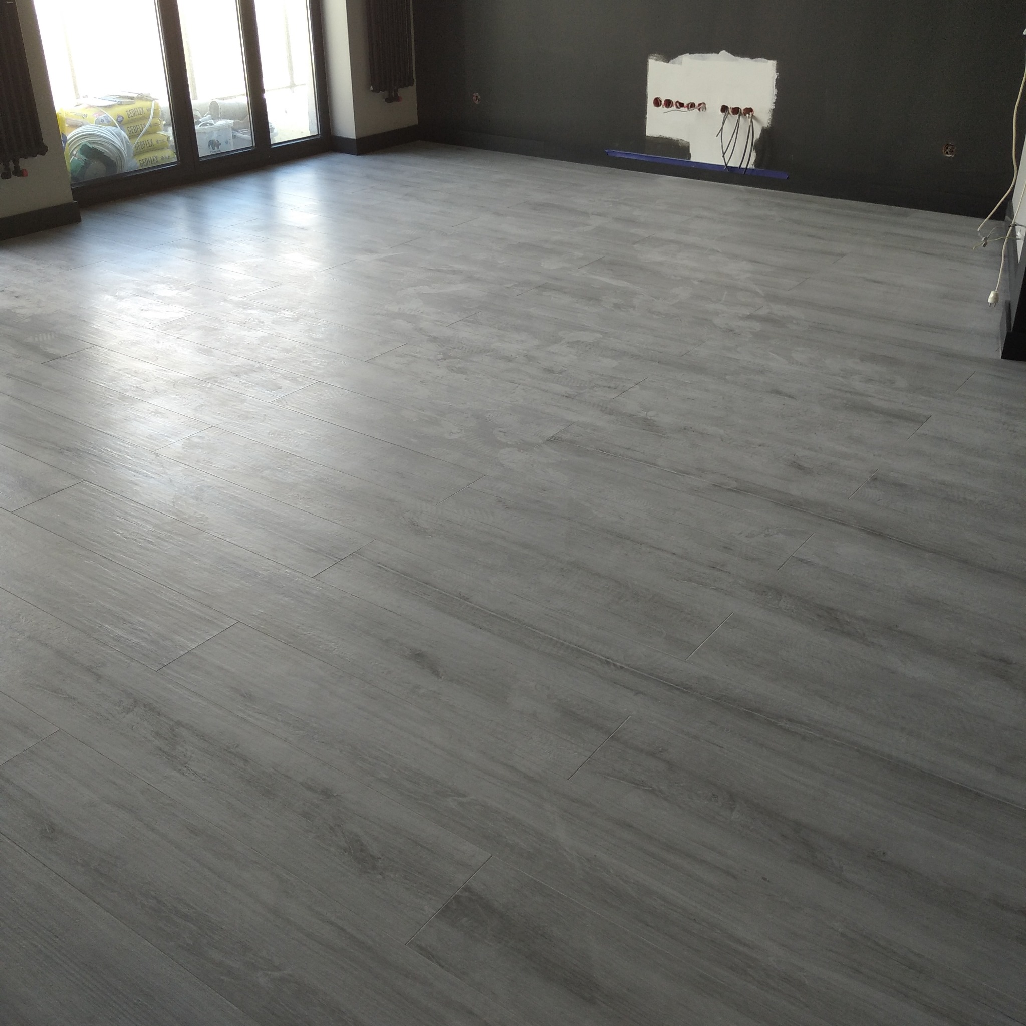 Ułożona podłoga z szarych, drewnopodobnych płytek w nowym pomieszczeniu z widocznymi instalacjami elektrycznymi w ścianie i workami z klejem za oknem.