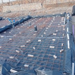 WIŚNIEWSKI DESIGN SPÓŁKA Z OGRANICZONĄ ODPOWIEDZIALNOŚCIĄ - Przygotowanie zbrojenia fundamentu na folii, z użyciem cegieł dystansowych, widoczne pionowe rury kanalizacyjne i laserowy niwelator, w tle skarpa zabezpieczona murem oporowym.