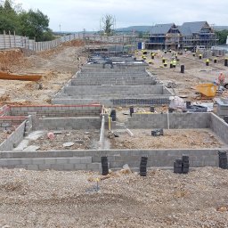 WIŚNIEWSKI DESIGN SPÓŁKA Z OGRANICZONĄ ODPOWIEDZIALNOŚCIĄ - Budowa fundamentów pod nowe budynki mieszkalne, widoczne bloczki betonowe, częściowo wymurowane ściany fundamentowe, wykopane rowy, koparka i pracownicy w tle.