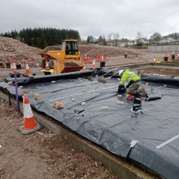 WIŚNIEWSKI DESIGN SPÓŁKA Z OGRANICZONĄ ODPOWIEDZIALNOŚCIĄ - Pracownik w odblaskowej kamizelce i kasku układa folię na przygotowanym fundamencie, w tle hałda gruzu i żółta wywrotka, teren budowy oznaczony pachołkami.