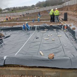 WIŚNIEWSKI DESIGN SPÓŁKA Z OGRANICZONĄ ODPOWIEDZIALNOŚCIĄ - Przygotowanie terenu pod wylewkę fundamentów: rozłożona czarna folia zabezpieczająca, drewniane klocki stabilizujące, widoczne zbrojenie i instalacje, pracownicy w odblaskowych kamizelkach w tle.