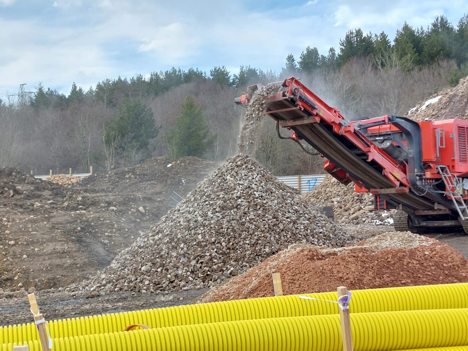 Czerwona kruszarka przesiewa kamienie, tworząc duży kopiec; w tle widać ziemię, drzewa i żółte rury.