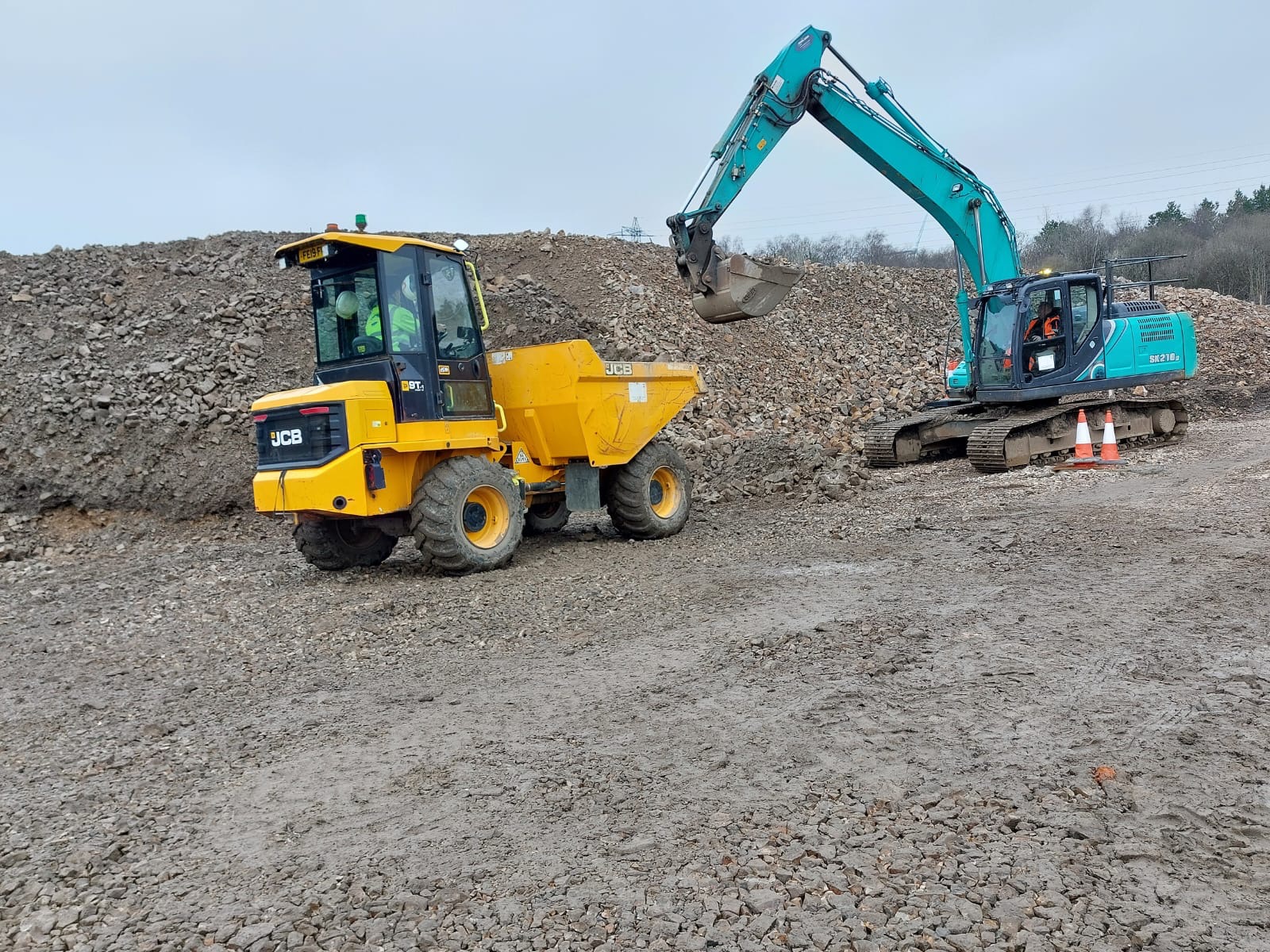 Żółta wywrotka JCB i turkusowa koparka Kobelco SK210LC na placu budowy z dużym stosem ziemi w tle, dwa pomarańczowe pachołki ostrzegawcze obok koparki.