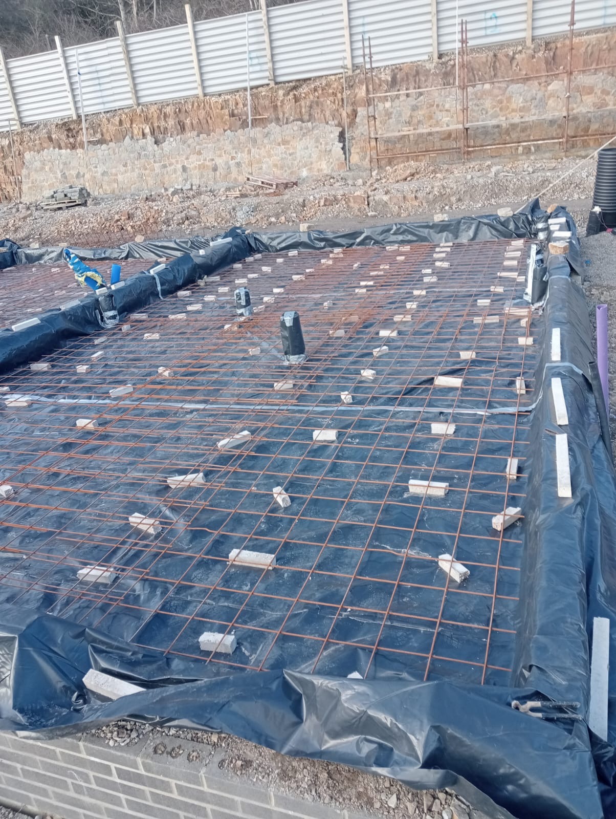 Przygotowanie zbrojenia fundamentu na folii, z użyciem cegieł dystansowych, widoczne pionowe rury kanalizacyjne i laserowy niwelator, w tle skarpa zabezpieczona murem oporowym.