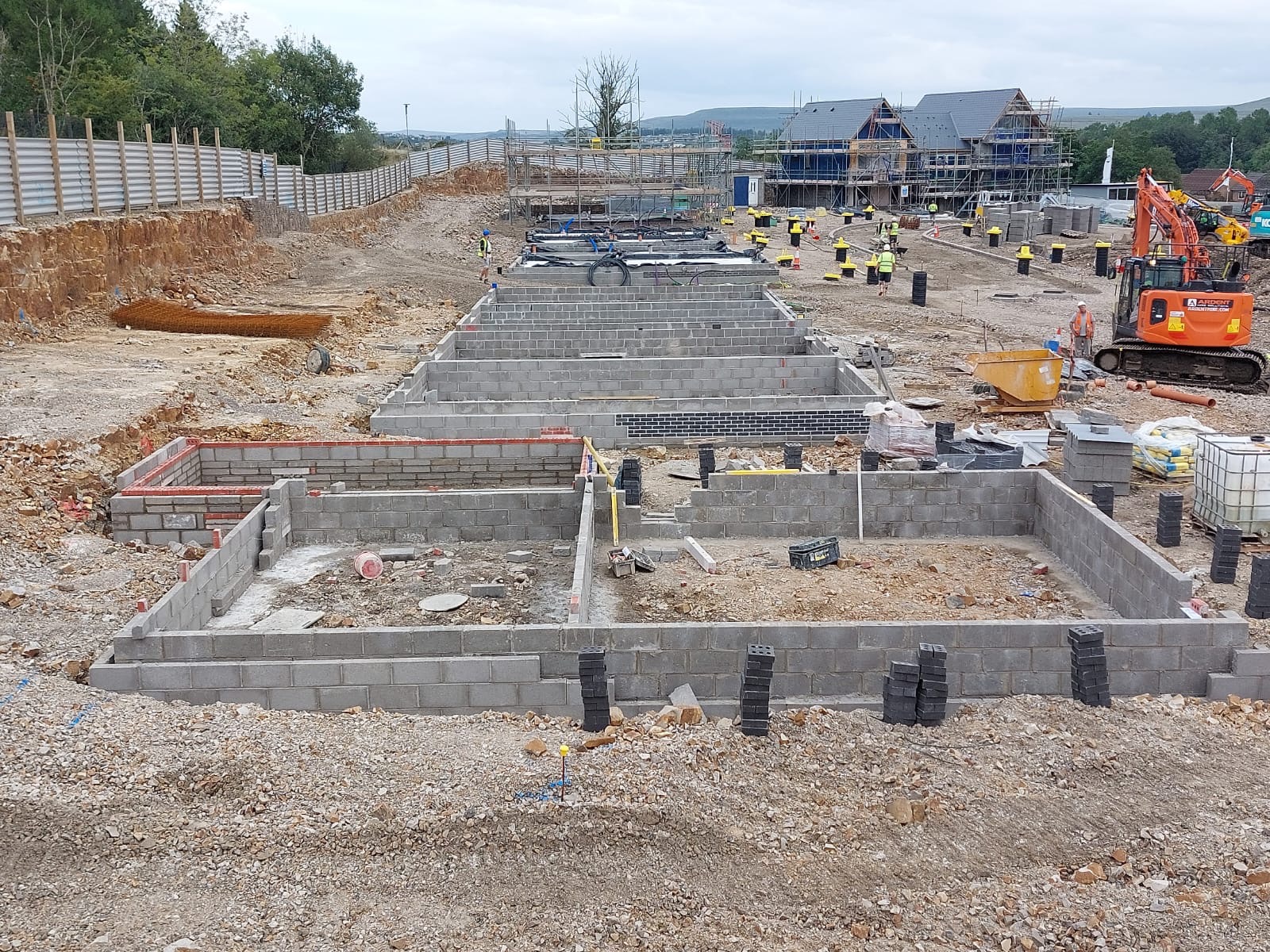 Budowa fundamentów pod nowe budynki mieszkalne, widoczne bloczki betonowe, częściowo wymurowane ściany fundamentowe, wykopane rowy, koparka i pracownicy w tle.