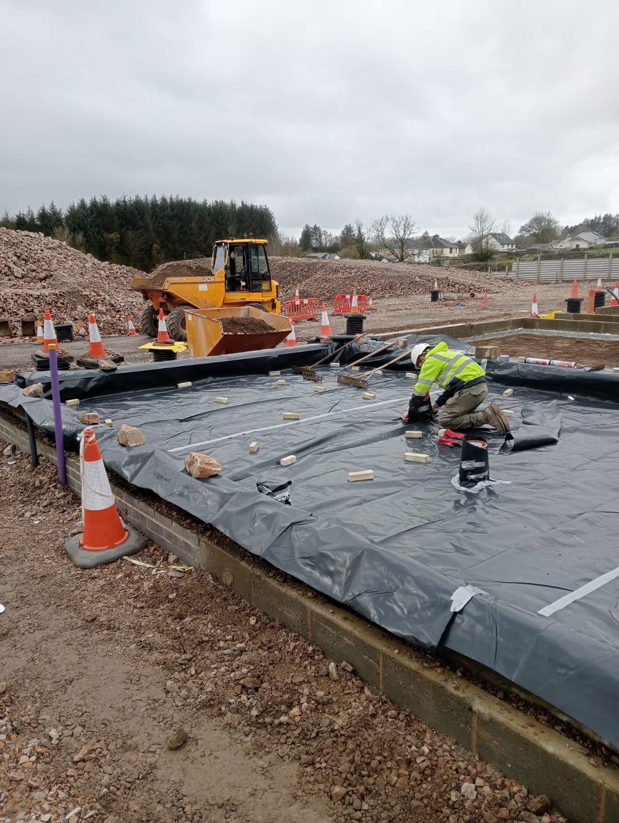 Pracownik w odblaskowej kamizelce i kasku układa folię na przygotowanym fundamencie, w tle hałda gruzu i żółta wywrotka, teren budowy oznaczony pachołkami.