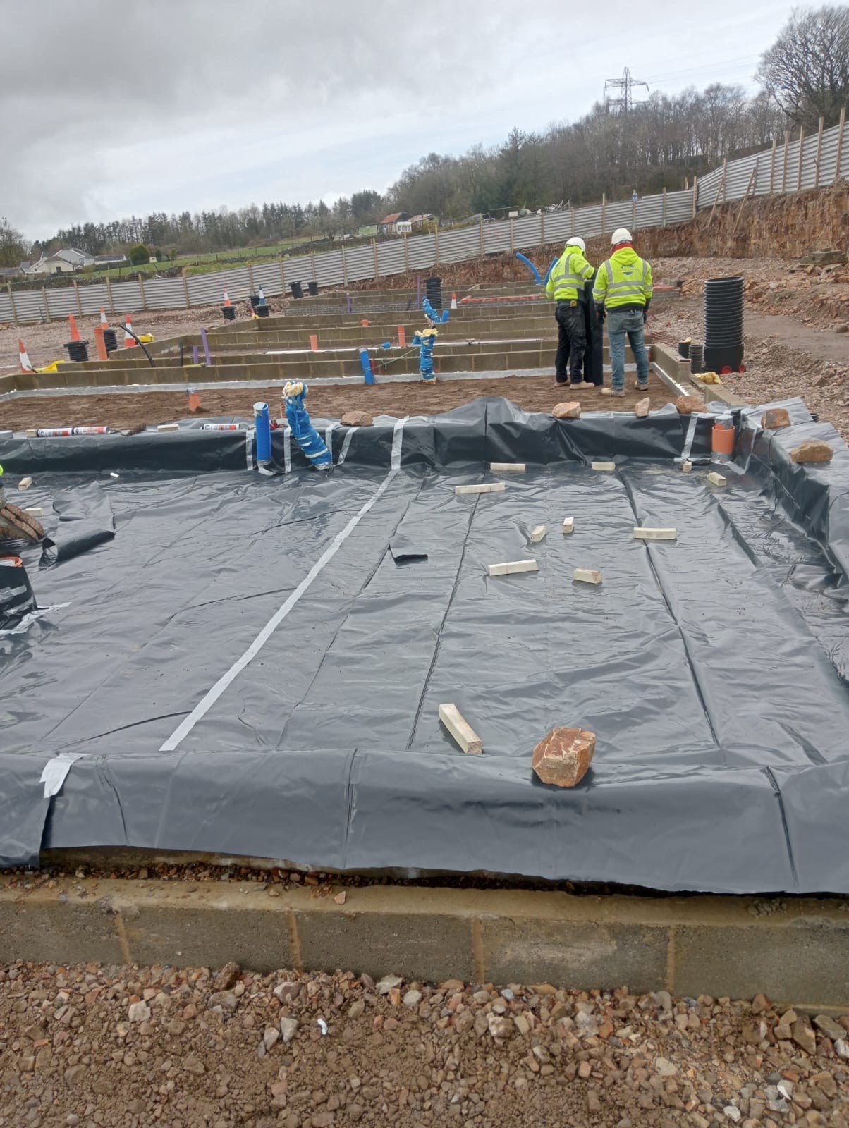 Przygotowanie terenu pod wylewkę fundamentów: rozłożona czarna folia zabezpieczająca, drewniane klocki stabilizujące, widoczne zbrojenie i instalacje, pracownicy w odblaskowych kamizelkach w tle.
