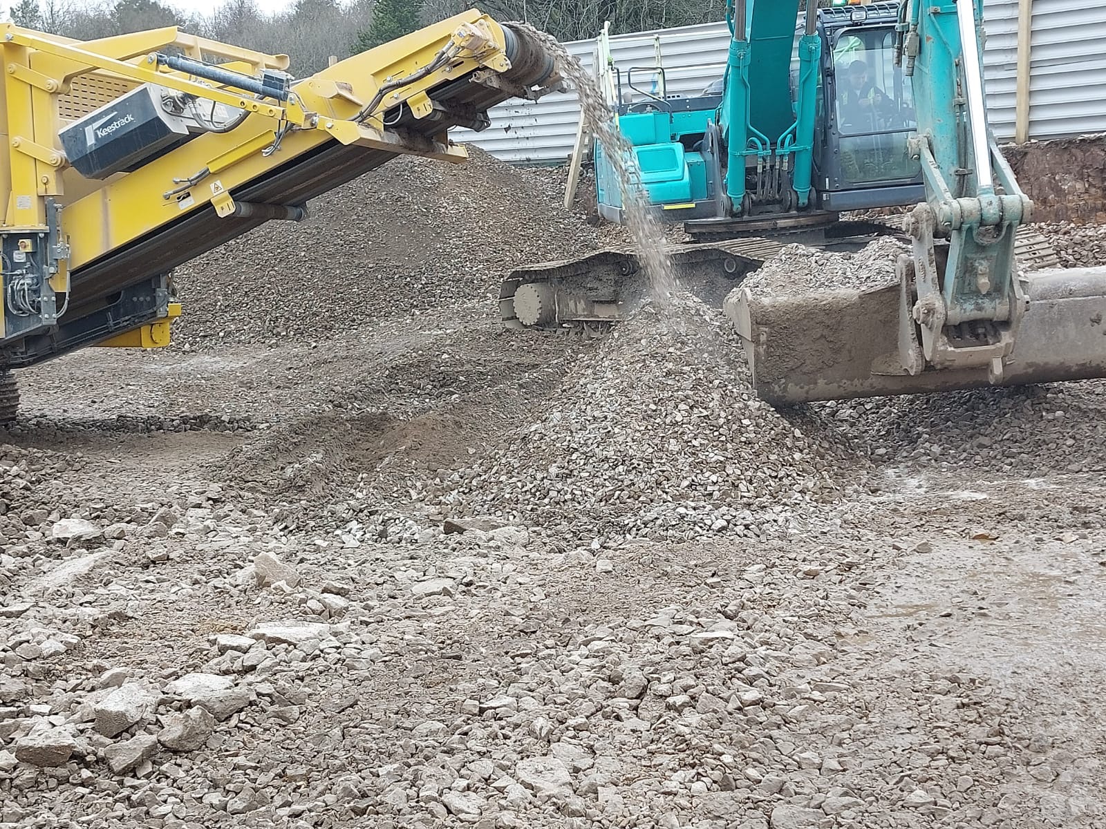 Żółty przesiewacz kruszywa Keestrack i niebieska koparka Hyundai, przesypywanie i układanie gruzu na placu budowy, widoczne ślady opon na ziemi.