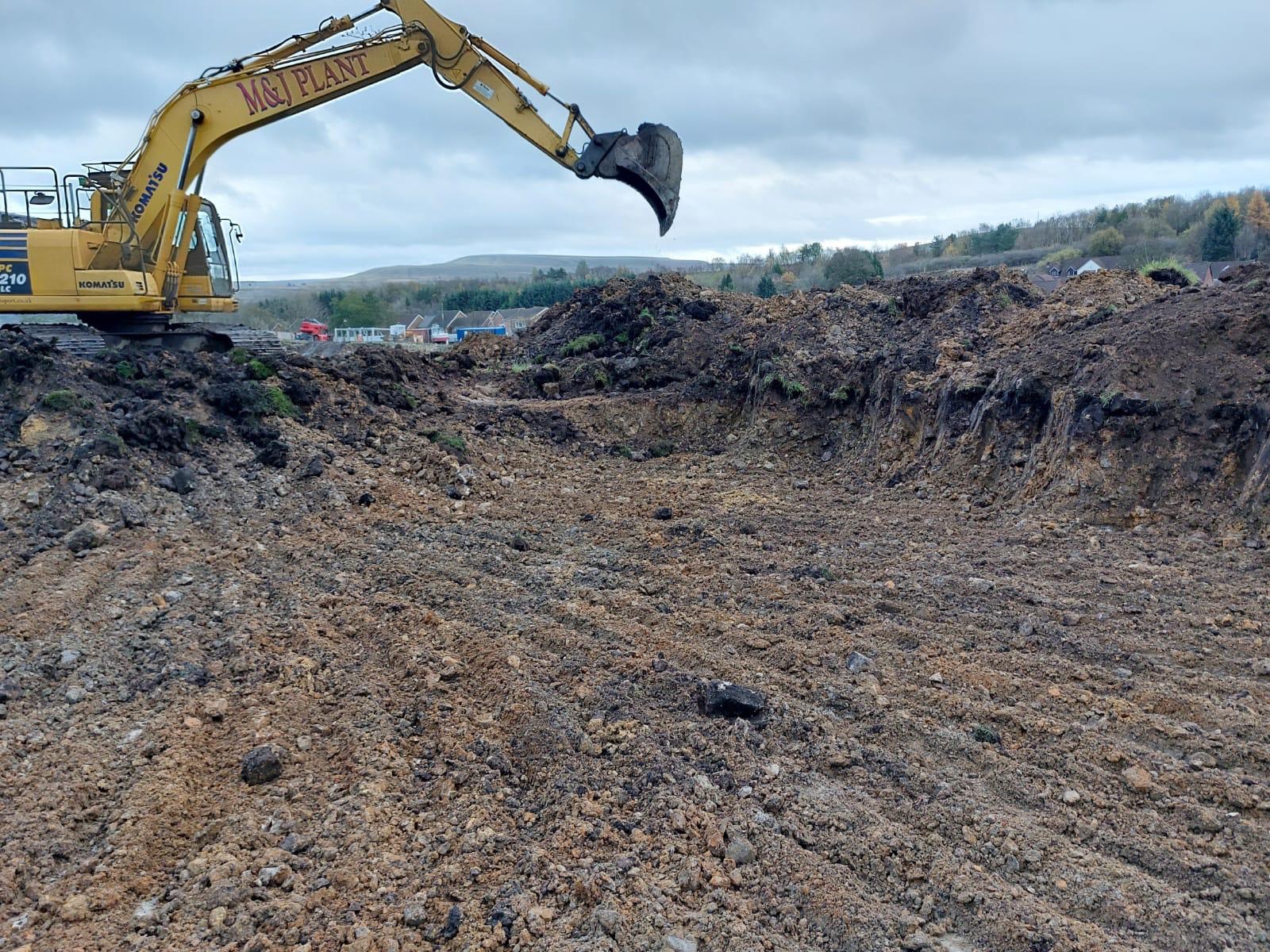 Żółta koparka Komatsu z łyżką unosi się nad wykopem w ziemi, w tle widoczne zabudowania i wzgórza pod pochmurnym niebem.