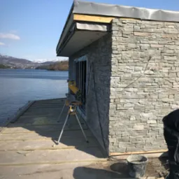 Budynek w trakcie wykończenia elewacji kamiennej na nabrzeżu, widoczna piła stołowa na trójnogu, w tle jezioro i góry.