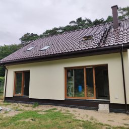 TREDOM BUDOWA DOMÓW SZKIELETOWYCH MAGDALENA SZWECH, SŁAWOMIR SZWECH SPÓŁKA JAWNA - Niedawno wybudowany dom jednorodzinny z brązową dachówką, brązowymi oknami i beżową elewacją, widoczne dwa okna dachowe i komin, teren wokół domu jeszcze niezagospodarowany.