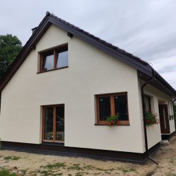 TREDOM BUDOWA DOMÓW SZKIELETOWYCH MAGDALENA SZWECH, SŁAWOMIR SZWECH SPÓŁKA JAWNA - Nowo wybudowany dom jednorodzinny z beżową elewacją, brązowym dachem i stolarką okienną w kolorze drewna, z małymi skrzynkami z czerwonymi kwiatami pod oknami.