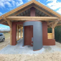 Zadaszona konstrukcja z cegły i drewna z elementami w kolorze szarym, prawdopodobnie grill lub piec, na utwardzonym placu z kostki brukowej, w tle samochód i ogrodzenie.