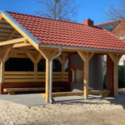 Drewniana altana z czerwonym dachem z dachówki ceramicznej, murowanym grillem i drewnianymi ławkami na kostce brukowej, otoczona piaszczystym terenem.