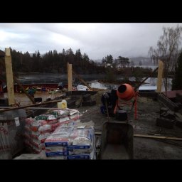 TREDOM BUDOWA DOMÓW SZKIELETOWYCH MAGDALENA SZWECH, SŁAWOMIR SZWECH SPÓŁKA JAWNA - Budowa domu murowanego w trakcie: widoczne fundamenty z bloczków betonowych, zapas cementu w workach, betoniarka i taczka na tle jeziora w pochmurny dzień.