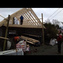 TREDOM BUDOWA DOMÓW SZKIELETOWYCH MAGDALENA SZWECH, SŁAWOMIR SZWECH SPÓŁKA JAWNA - Konstrukcja drewnianego szkieletu dachu na murowanym budynku gospodarczym, widoczny pracownik na dachu oraz materiały budowlane na ziemi.