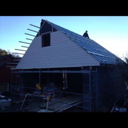 TREDOM BUDOWA DOMÓW SZKIELETOWYCH MAGDALENA SZWECH, SŁAWOMIR SZWECH SPÓŁKA JAWNA - Wznoszenie konstrukcji domu z widocznym fragmentem dachu pokrytego folią, ścianą szczytową z oknem i rusztowaniami na tle błękitnego nieba.