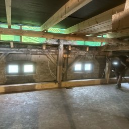 TREDOM BUDOWA DOMÓW SZKIELETOWYCH MAGDALENA SZWECH, SŁAWOMIR SZWECH SPÓŁKA JAWNA - Wnętrze budynku w trakcie prac budowlanych: widoczne drewniane belki stropowe, fragment ściany z dwoma małymi oknami, podłoga z płyt OSB i pracownik sprzątający pomieszczenie miotłą.