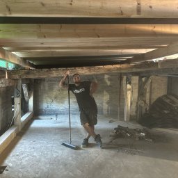 TREDOM BUDOWA DOMÓW SZKIELETOWYCH MAGDALENA SZWECH, SŁAWOMIR SZWECH SPÓŁKA JAWNA - Uśmiechnięty mężczyzna oparty o miotłę w trakcie prac przygotowawczych do remontu poddasza, widoczne nowe drewniane belki stropowe i fragment starej ceglanej ściany.