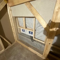 TREDOM BUDOWA DOMÓW SZKIELETOWYCH MAGDALENA SZWECH, SŁAWOMIR SZWECH SPÓŁKA JAWNA - Wnętrze w Gdyni w trakcie budowy domu szkieletowego, widoczne drewniane elementy konstrukcyjne, okno z folią ochronną i fragment ściany z płyt kartonowo-gipsowych.