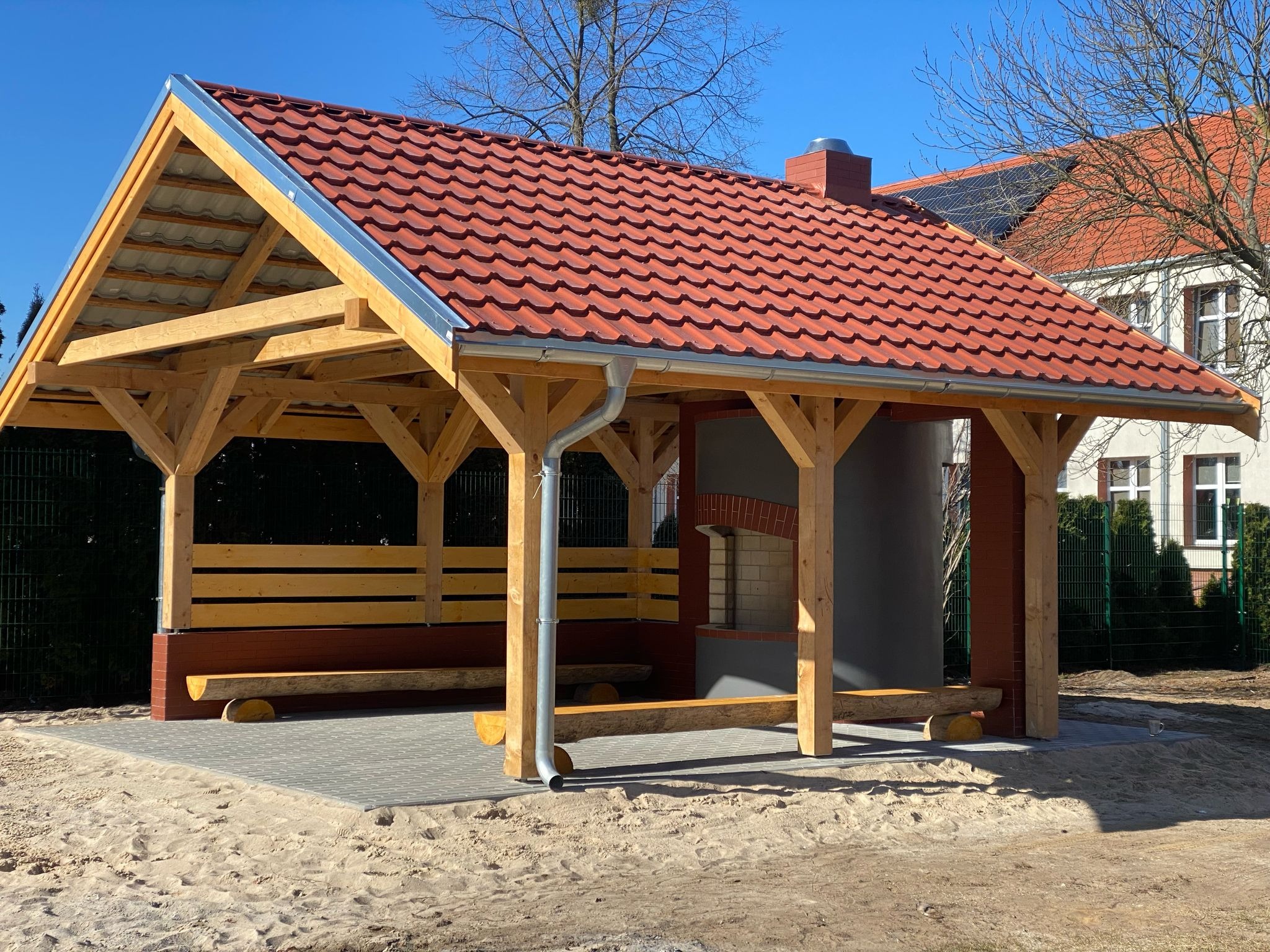 Drewniana altana z czerwonym dachem z dachówki ceramicznej, murowanym grillem i drewnianymi ławkami na kostce brukowej, otoczona piaszczystym terenem.