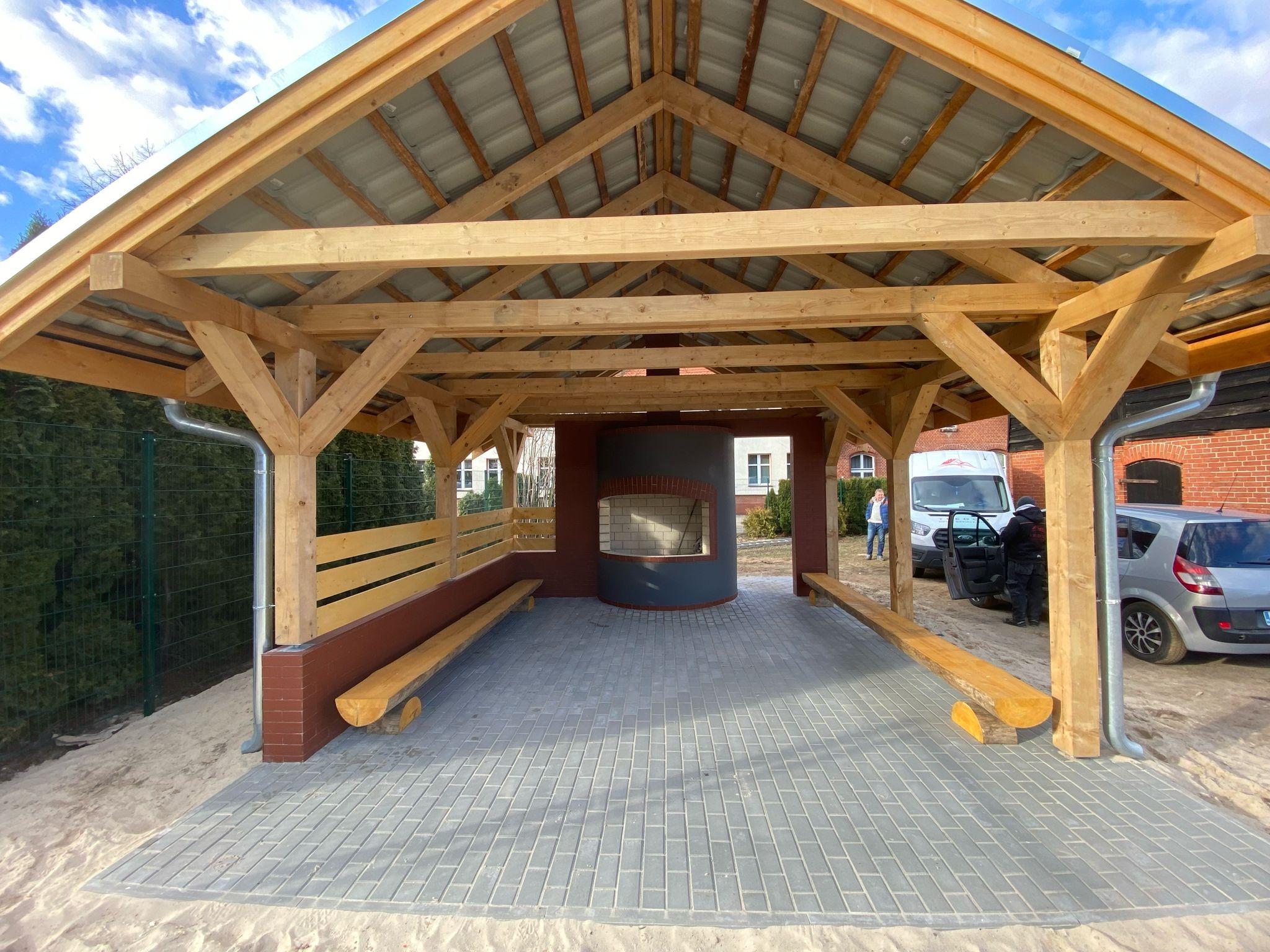 Zadaszona altana z drewnianą konstrukcją, murowanym grillem i ławkami w Gdyni, wyłożona szarą kostką brukową, otoczona piaskiem.