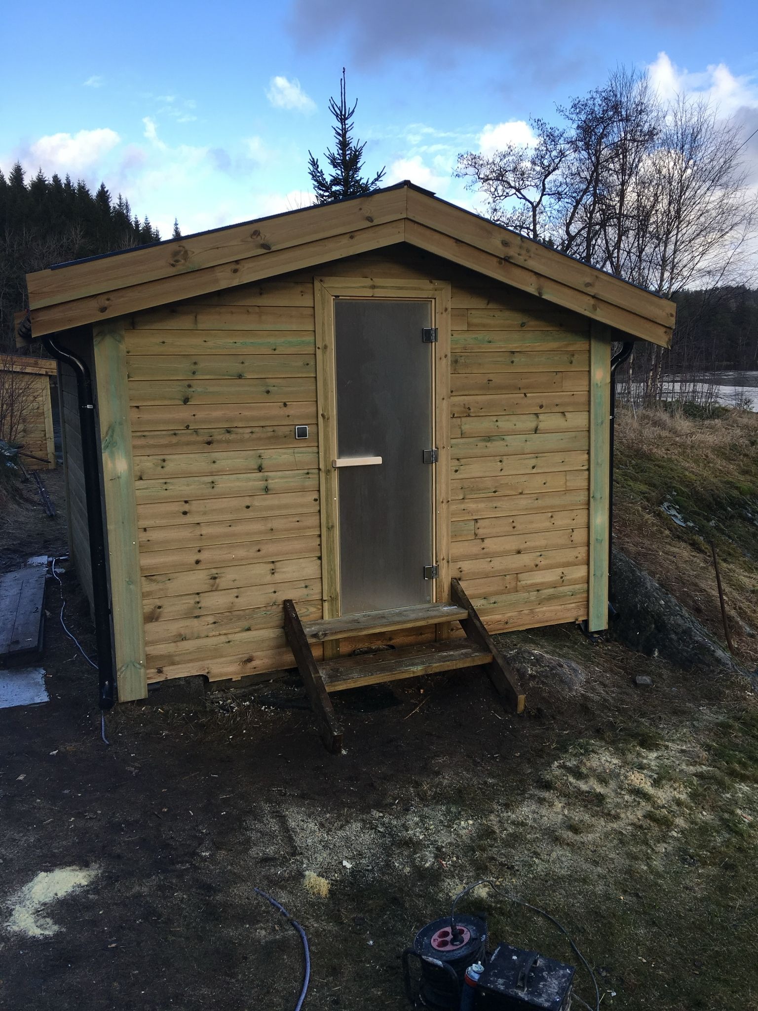 Nowo wybudowany drewniany domek z przeszklonymi drzwiami i schodkami, umieszczony na wzniesieniu porośniętym trawą i mchem, w tle drzewa i częściowo zamarznięte jezioro.