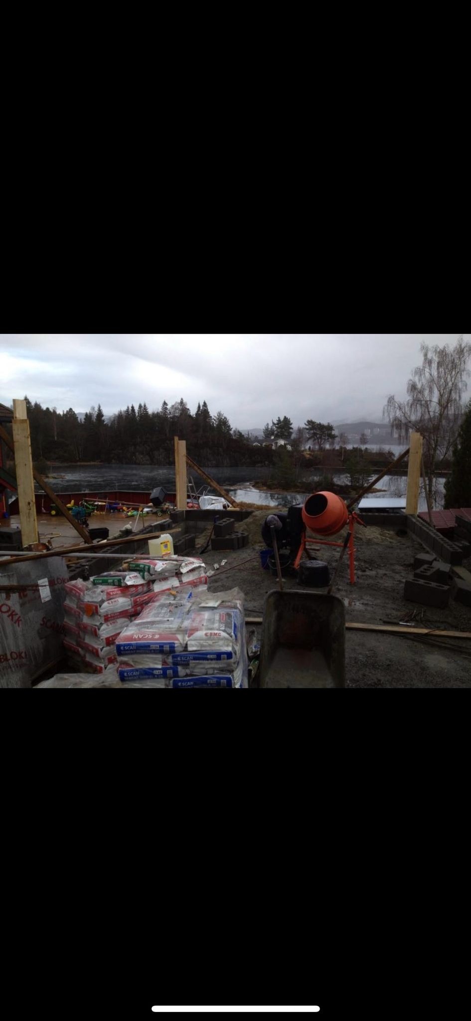 Budowa domu murowanego w trakcie: widoczne fundamenty z bloczków betonowych, zapas cementu w workach, betoniarka i taczka na tle jeziora w pochmurny dzień.