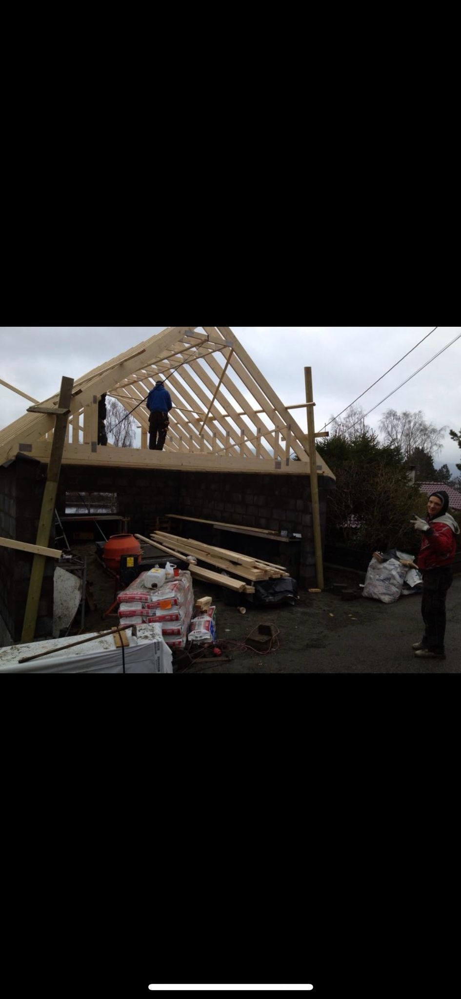 Konstrukcja drewnianego szkieletu dachu na murowanym budynku gospodarczym, widoczny pracownik na dachu oraz materiały budowlane na ziemi.