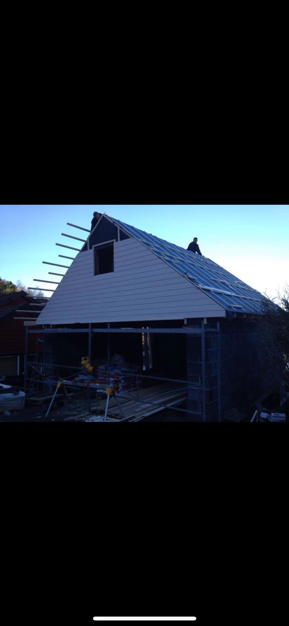 Wznoszenie konstrukcji domu z widocznym fragmentem dachu pokrytego folią, ścianą szczytową z oknem i rusztowaniami na tle błękitnego nieba.