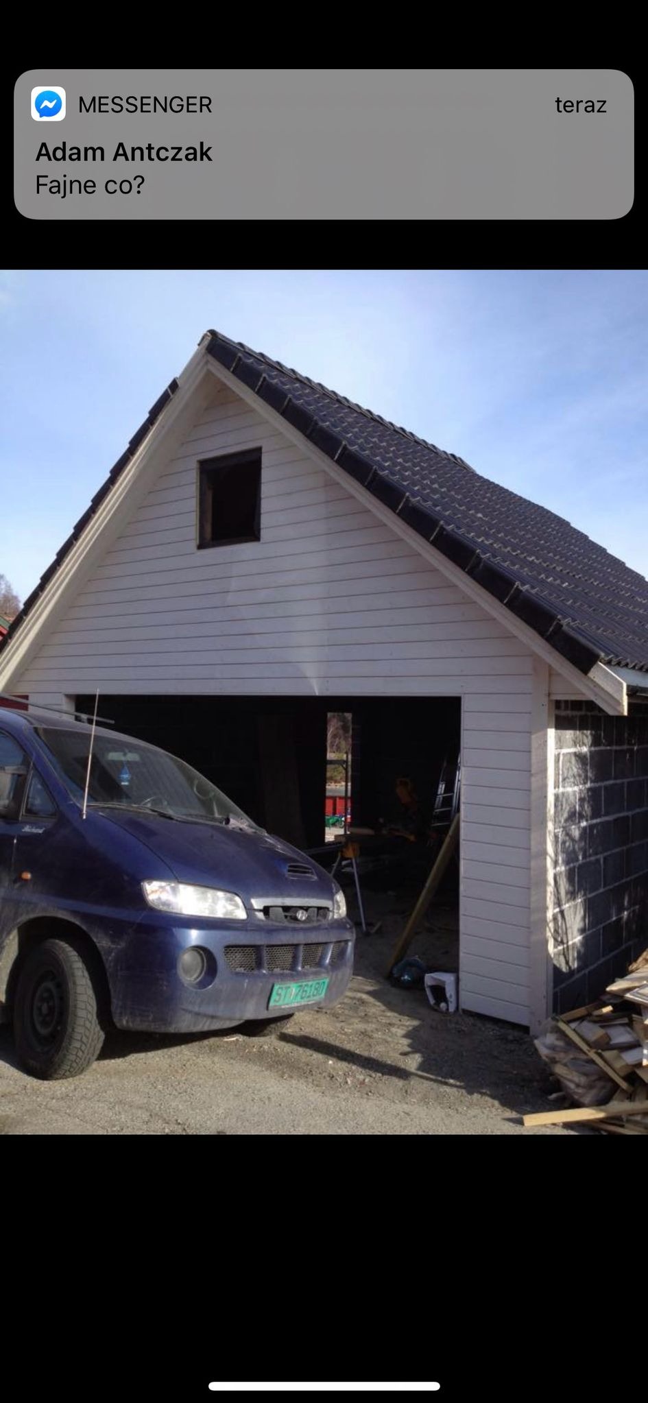 Budowa garażu z białymi panelami elewacyjnymi i czarnym dachem, widoczny fragment murowanej ściany z szarego bloczka oraz niebieski samochód na pierwszym planie.
