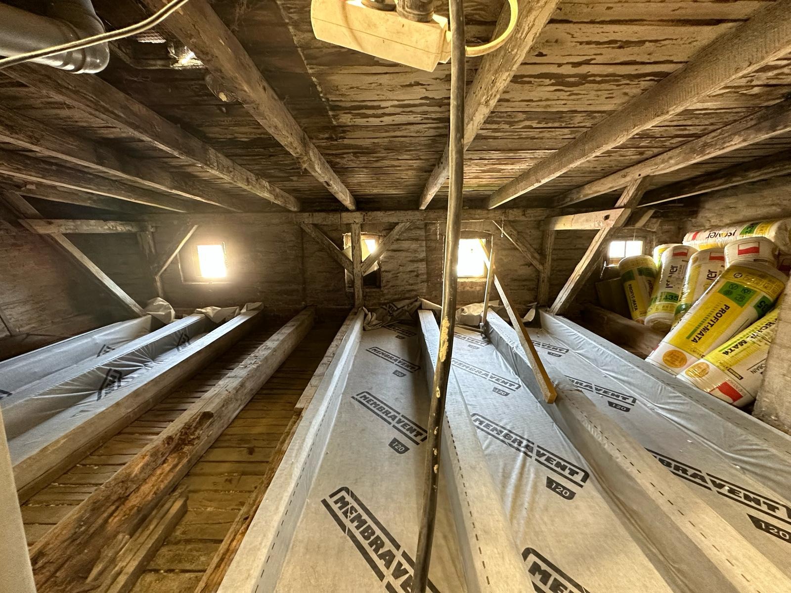 Strych z widocznymi krokwiami pokrytymi membraną dachową Membravent 120, w tle materiały izolacyjne Profit-Mata i małe okna dachowe.