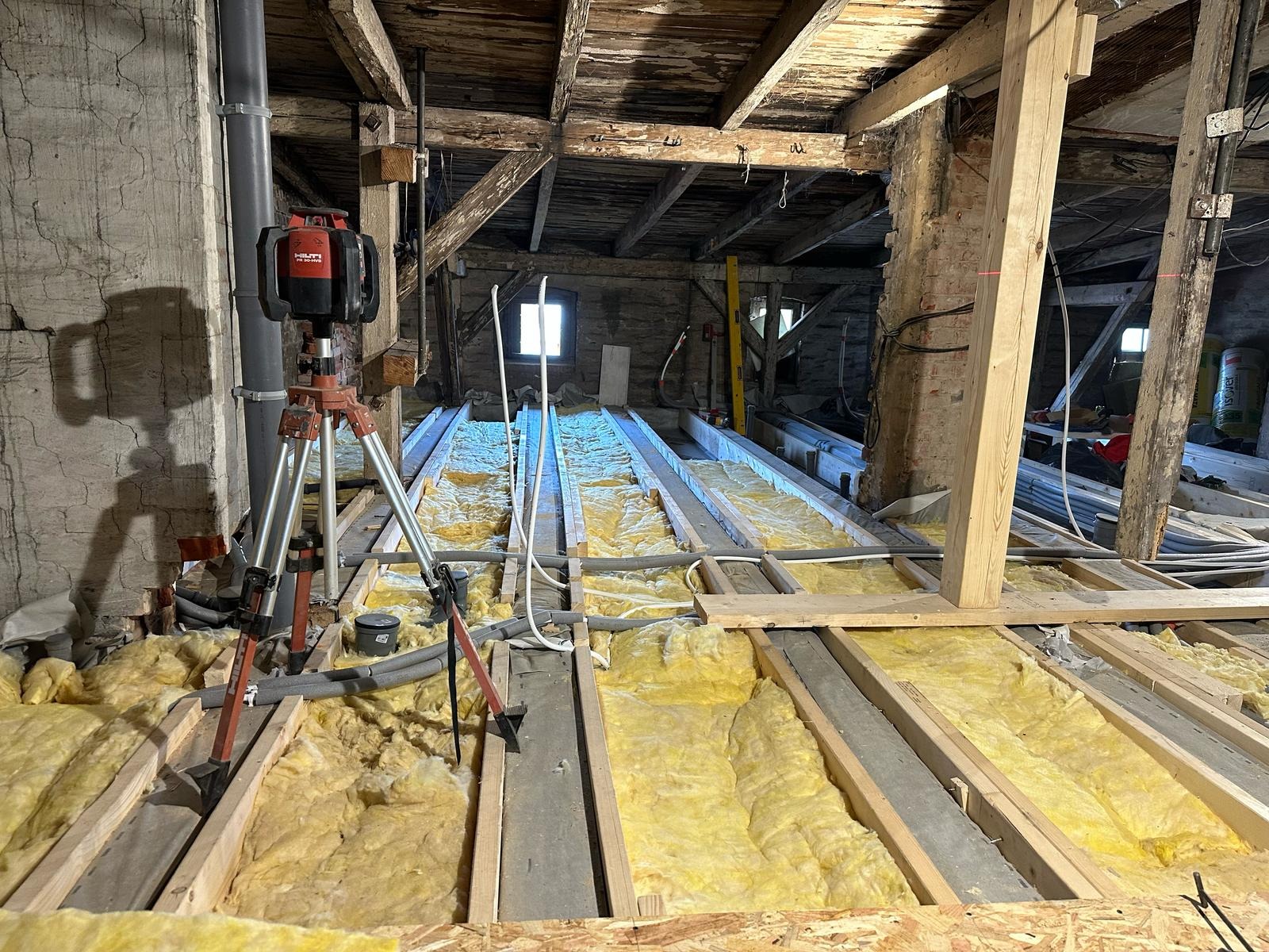 Montaż izolacji z wełny mineralnej między legarami na poddaszu, widoczny laser krzyżowy Hilti na statywie, stare drewniane krokwie i ceglana ściana w tle