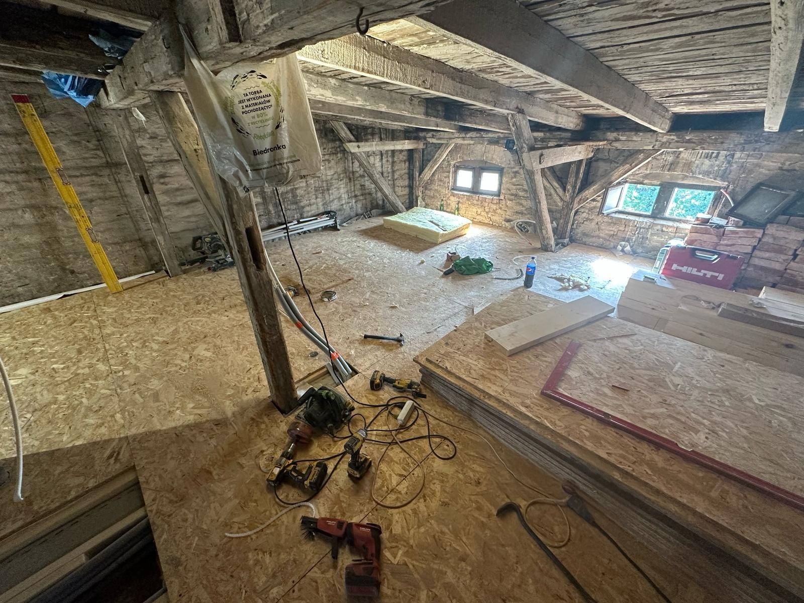 Adaptacja poddasza: surowe drewniane belki, ceglane ściany, podłoga wyłożona płytami OSB, narzędzia budowlane i materiały izolacyjne w worku foliowym z logotypem sklepu spożywczego.