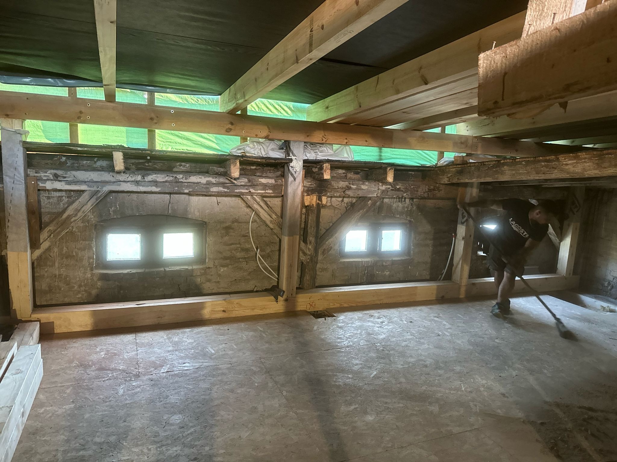 Wnętrze budynku w trakcie prac budowlanych: widoczne drewniane belki stropowe, fragment ściany z dwoma małymi oknami, podłoga z płyt OSB i pracownik sprzątający pomieszczenie miotłą.